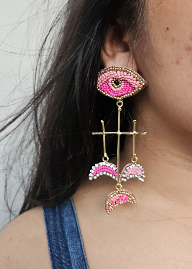 Close-up of a person wearing pink and gold earrings with a neutral background evil eye