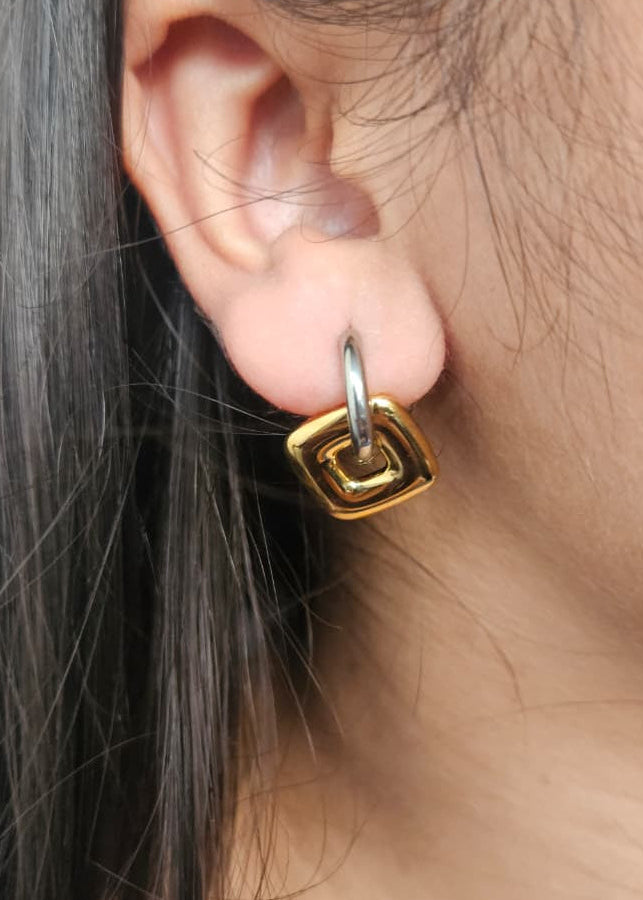 Gold square earring worn by a person with dark hair.