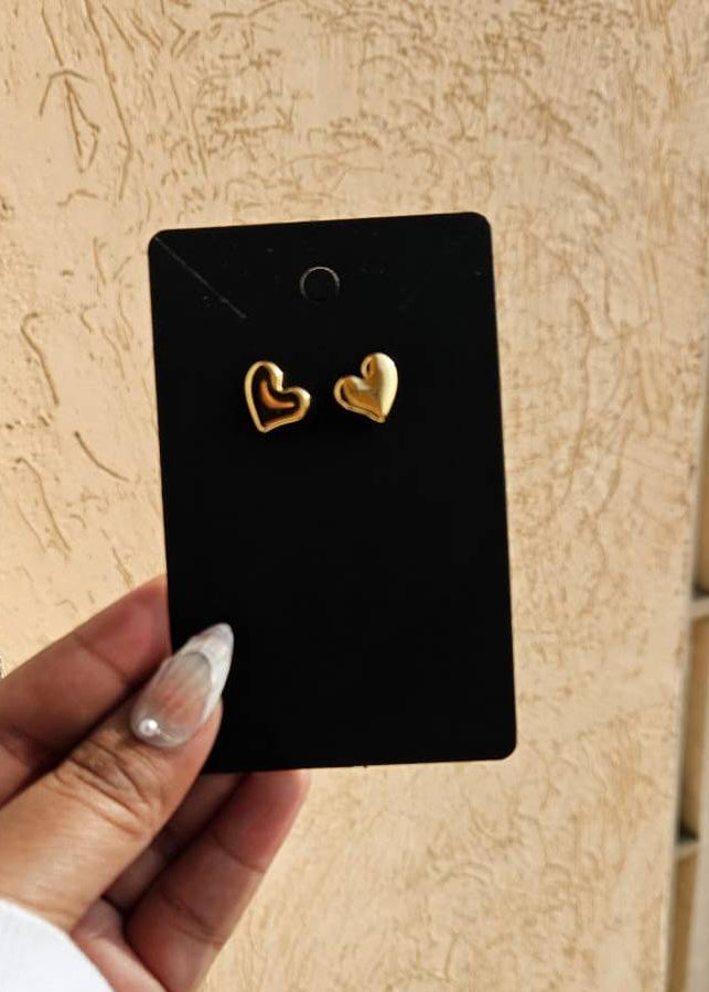 Gold heart-shaped earrings on a black card held by a hand against a beige wall.