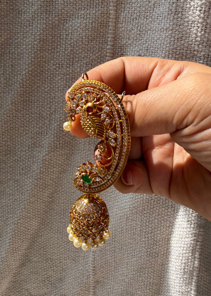 A hand holding a detailed gold-toned traditional Indian jhumka earring with intricate stone work, a peacock motif, and a dangling bell-shaped charm, set against a textured gray fabric background.