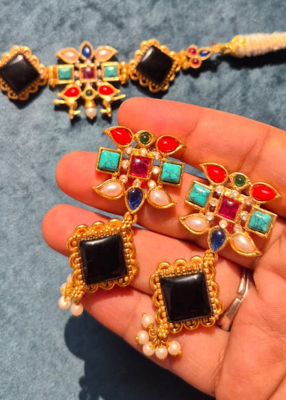 Colorful jewelry set with gold frames, black stones, and multicolored gemstones on a hand against a blue background.