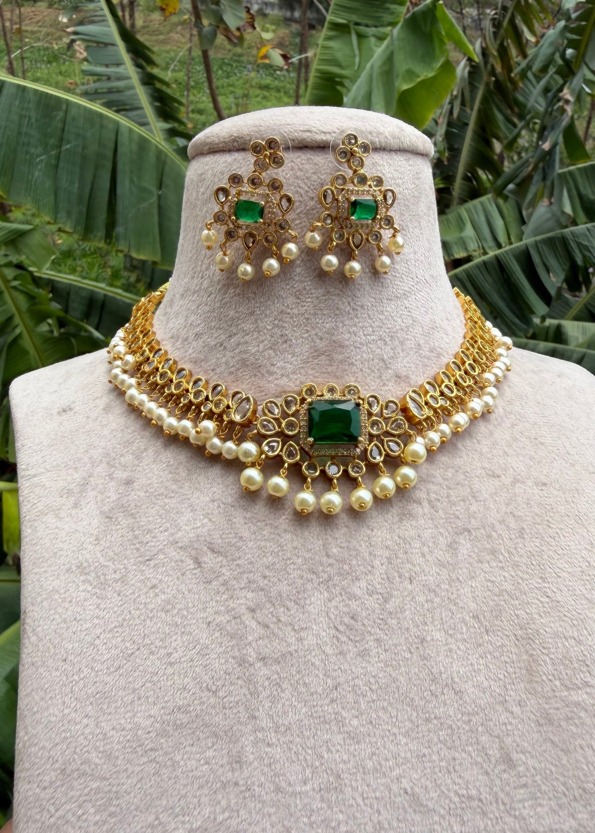Gold and green stone jewelry set displayed on a beige velvet mannequin bust, featuring a choker necklace and matching earrings, with a tropical garden background.