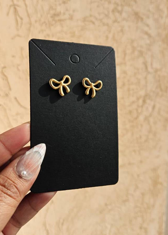 Gold bow-shaped earrings on a black card held by a hand against a beige background