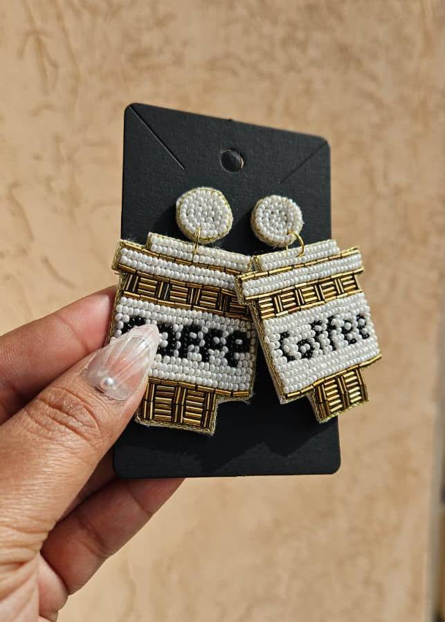 Pair of earrings shaped like coffee bags with 'coffee' text on a black card, held by a hand against a beige background.