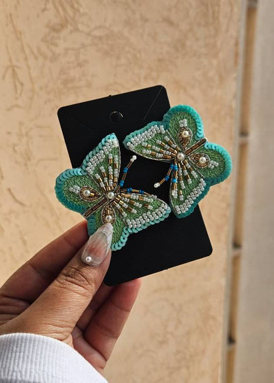 Butterfly-shaped earrings with green and gold details on a black card, held by a hand.