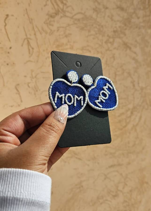 Blue heart-shaped earrings with 'MOM' text held by a hand on a beige background