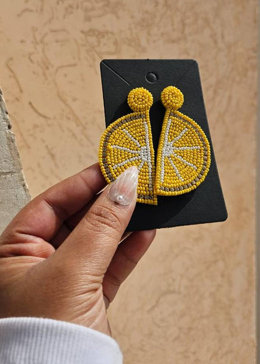 Hand holding a pair of yellow beaded earrings on a black card against a beige wall.