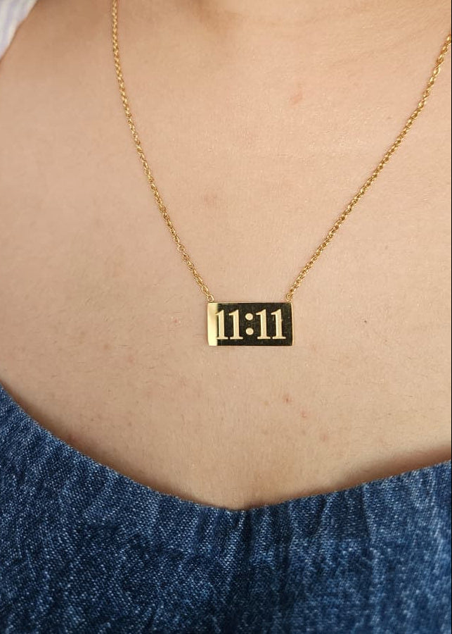 Gold necklace with a rectangular pendant displaying '11:11' worn by a person against a blue denim background.