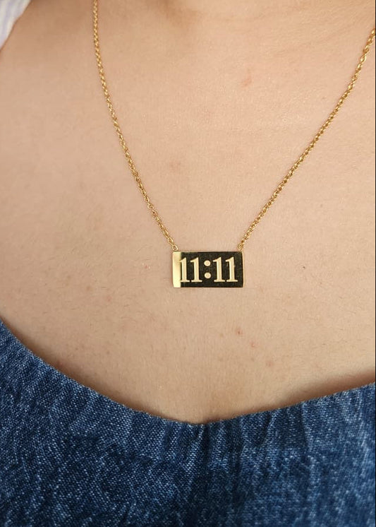 Gold necklace with a rectangular pendant displaying '11:11' worn by a person against a blue denim background.
